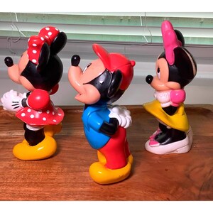 Pu&ograve; includere: Tre colorate statuette di Topolino e Minnie. Topolino indossa un cappello rosso, una camicia blu e pantaloni rossi. Minnie indossa un vestito rosso a pois e un fiocco. Le statuette sono su una superficie di legno.