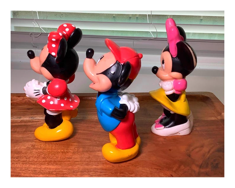 Pu&ograve; includere: Tre colorate statuette di Topolino e Minnie. Topolino indossa un cappello rosso, una camicia blu e pantaloni rossi. Minnie indossa un vestito rosso a pois e un fiocco. Le statuette sono su una superficie di legno.