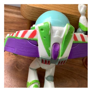May include: A Buzz Lightyear action figure, with a light blue helmet, white body, and green and purple details. The figure has a jetpack with red and white stripes and green wings. The toy is a collectible item.