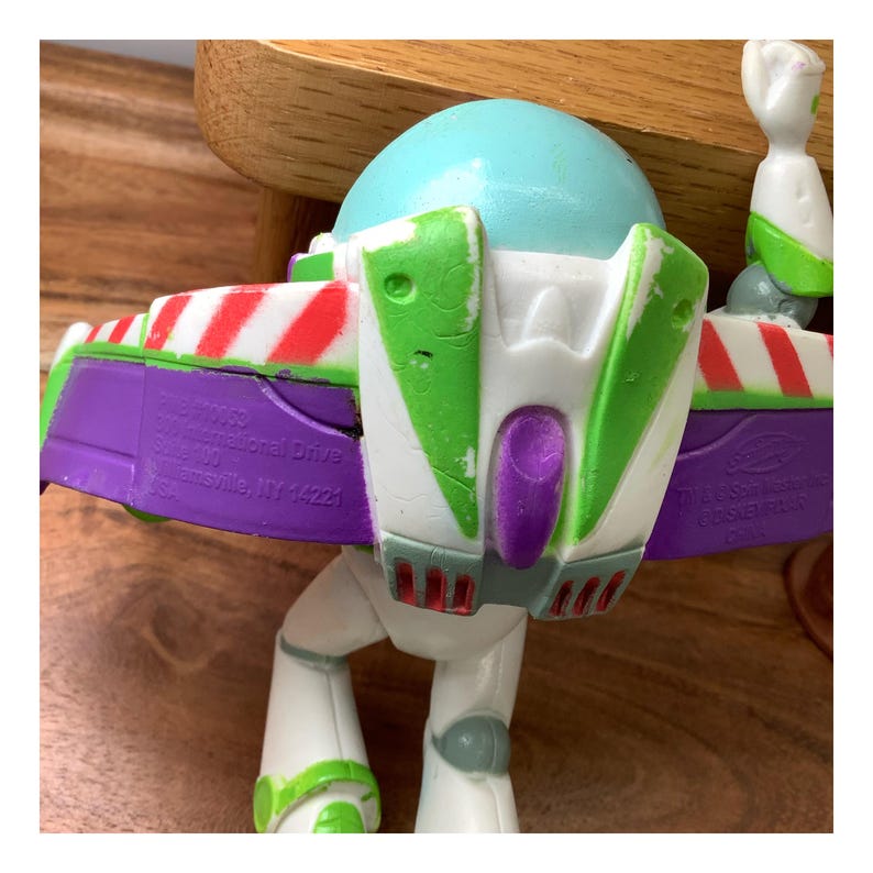May include: A Buzz Lightyear action figure, with a light blue helmet, white body, and green and purple details. The figure has a jetpack with red and white stripes and green wings. The toy is a collectible item.