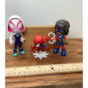 Op de afbeelding: Drie superheldenbeeldjes: Spider-Gwen, Spider-Man en Ms. Marvel. Spider-Gwen draagt een witte en roze capuchon met een paars en wit pak. Spider-Man is rood en blauw. Ms. Marvel draagt een blauw en goud pak. Een houten liniaal staat op de voorgrond.