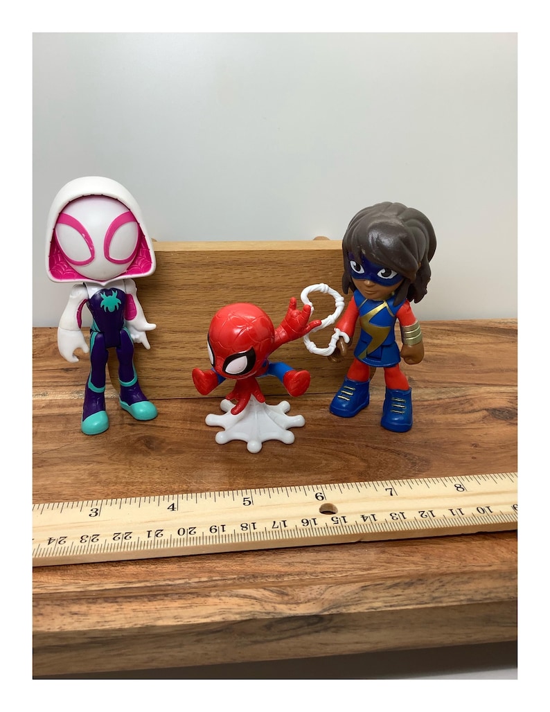 Op de afbeelding: Drie superheldenbeeldjes: Spider-Gwen, Spider-Man en Ms. Marvel. Spider-Gwen draagt een witte en roze capuchon met een paars en wit pak. Spider-Man is rood en blauw. Ms. Marvel draagt een blauw en goud pak. Een houten liniaal staat op de voorgrond.