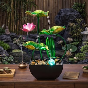 以下が含まれることがあります： 蓮の花と葉のデザインの装飾的な卓上噴水。噴水は、ピンクの蓮の花、緑と黄色のスイレンの葉、そして輝く青いオーブが特徴です。水はスイレンの葉から小石で満たされた黒いボウルに流れ落ちます。