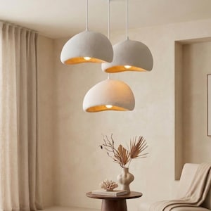 Puede incluir: Tres lámparas colgantes con un exterior texturizado blanquecino y un interior dorado y cálido. Las luces cuelgan de cables blancos contra una pared de tonos neutros. La habitación está decorada con una mesa pequeña, una silla y una ventana con una cortina beige.