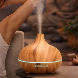 Puede incluir: Un difusor de aceites esenciales de madera con una línea de acento azul claro y un pequeño panel de control. El difusor está sobre una pequeña mesa negra. El vapor se eleva desde la parte superior del difusor. Una mujer se relaja con los brazos detrás de la cabeza.