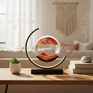 K&ouml;nnte beinhalten: Eine moderne Sandkunstlampe mit schwarzem Sockel und kreisf&ouml;rmigem Rahmen. Im Inneren der klaren Glaskugel bildet oranger Sand eine dynamische Landschaft. Die Lampe steht auf einem Holztisch, mit einer kleinen Sukkulente und B&uuml;chern in der N&auml;he.
