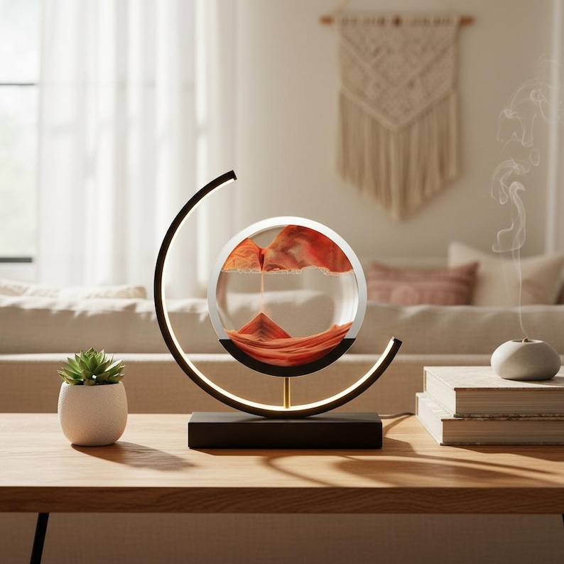 K&ouml;nnte beinhalten: Eine moderne Sandkunstlampe mit schwarzem Sockel und kreisf&ouml;rmigem Rahmen. Im Inneren der klaren Glaskugel bildet oranger Sand eine dynamische Landschaft. Die Lampe steht auf einem Holztisch, mit einer kleinen Sukkulente und B&uuml;chern in der N&auml;he.