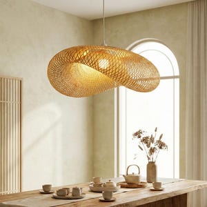 May include: A woven, golden pendant light hangs above a wooden dining table. The light has an organic shape and casts a warm glow. The dining table is set with a tea service and a vase of dried flowers. The light is made of woven bamboo.