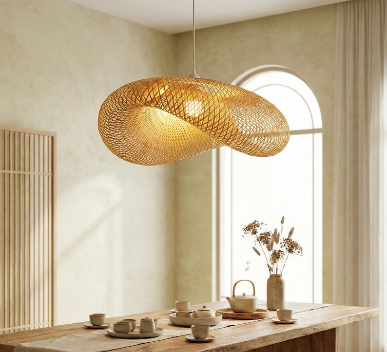 May include: A woven, golden pendant light hangs above a wooden dining table. The light has an organic shape and casts a warm glow. The dining table is set with a tea service and a vase of dried flowers. The light is made of woven bamboo.