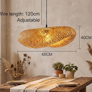 May include: A woven rattan pendant light, 60cm long and 40cm high, hangs above a wooden table. The light has a warm, golden colour and is lit from within. The wire is 120cm long and adjustable. The table is set with plates and plants.
