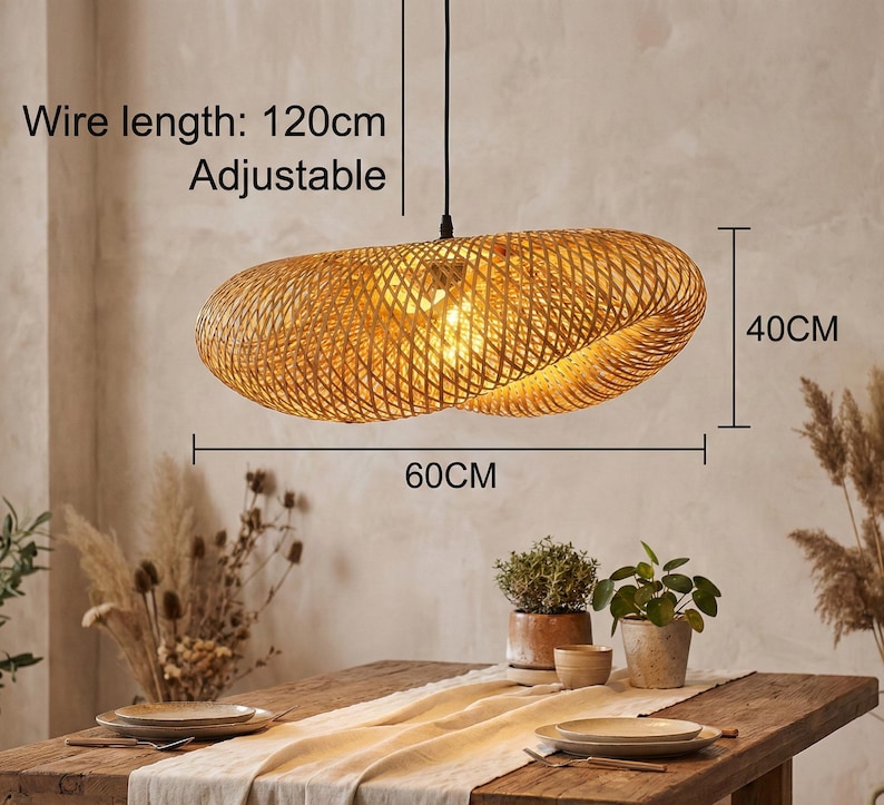 May include: A woven rattan pendant light, 60cm long and 40cm high, hangs above a wooden table. The light has a warm, golden colour and is lit from within. The wire is 120cm long and adjustable. The table is set with plates and plants.