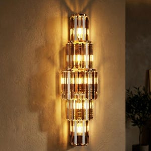 Può includere: Applique da parete verticale a più livelli con una calda luce ambrata. L'apparecchio è composto da elementi di cristallo rettangolari impilati, creando un design verticale. La sorgente luminosa è visibile, proiettando una luce soffusa.