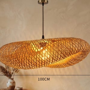 May include: A woven rattan pendant light. The oval-shaped light fitting is 100cm long and 55cm wide. It hangs from a black cord and a brass-coloured ceiling mount. The light is illuminated from within.