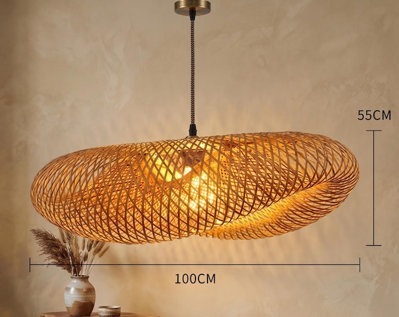 May include: A woven rattan pendant light. The oval-shaped light fitting is 100cm long and 55cm wide. It hangs from a black cord and a brass-coloured ceiling mount. The light is illuminated from within.