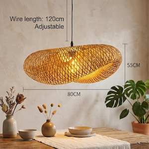 May include: A woven, oval pendant light. The light fitting is made of natural-coloured material and is 80cm long and 55cm wide. The wire length is 120cm and adjustable. The light hangs above a wooden table.
