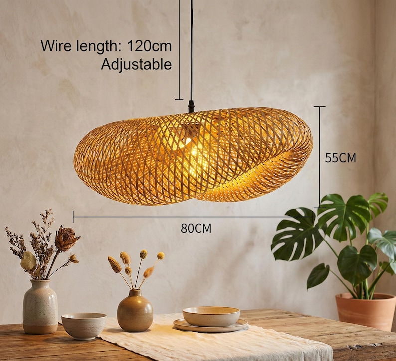 May include: A woven, oval pendant light. The light fitting is made of natural-coloured material and is 80cm long and 55cm wide. The wire length is 120cm and adjustable. The light hangs above a wooden table.