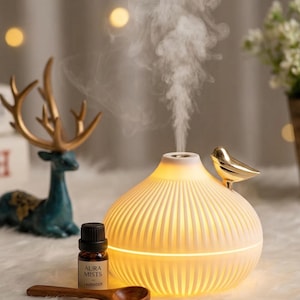 May include: A glowing, white ceramic essential oil diffuser with a gold bird accent. The diffuser is emitting steam, and a bottle of lavender oil and a wooden spoon are nearby. The text "Aura Mists" is visible on the oil bottle.