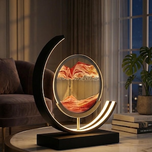 K&ouml;nnte beinhalten: Eine moderne dekorative Sandkunst-Lampe. Die Lampe hat einen schwarzen Sockel und Rahmen mit einem kreisf&ouml;rmigen Glaspaneel, das geschichteten Sand in Rot-, Orange- und Goldt&ouml;nen enth&auml;lt. Der Rahmen hat ein geschwungenes Design mit integrierter warmwei&szlig;er LED-Beleuchtung.