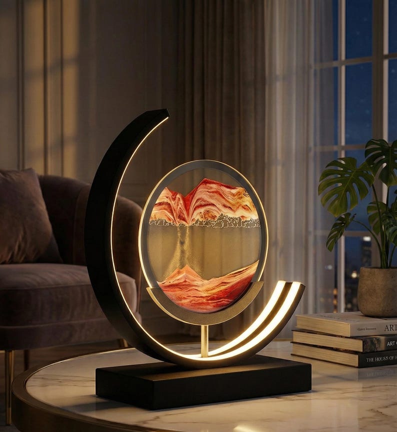 K&ouml;nnte beinhalten: Eine moderne dekorative Sandkunst-Lampe. Die Lampe hat einen schwarzen Sockel und Rahmen mit einem kreisf&ouml;rmigen Glaspaneel, das geschichteten Sand in Rot-, Orange- und Goldt&ouml;nen enth&auml;lt. Der Rahmen hat ein geschwungenes Design mit integrierter warmwei&szlig;er LED-Beleuchtung.