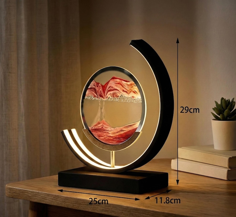 K&ouml;nnte beinhalten: Dekorative Sandkunst-Lampe mit schwarzem Sockel und halbmondf&ouml;rmigem Rahmen. Die Lampe verf&uuml;gt &uuml;ber ein kreisf&ouml;rmiges Glasgeh&auml;use mit rotem und wei&szlig;em Sand, das 29 cm hoch, 25 cm breit und 11,8 cm tief ist.