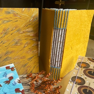 Può includere: Due diari fatti a mano con copertine gialle. Un diario ha un motivo di spighe di grano e l'altro ha una copertina gialla liscia con cuciture blu. Entrambi i diari sono chiusi e sono su un tavolo.