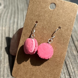 Könnte beinhalten: Ein Paar rosa Macaron-Ohrringe mit silberfarbenen Haken. Die Ohrringe werden auf einem braunen Kartonanhänger präsentiert. Die Macarons haben ein strukturiertes Aussehen mit einem genähten Design an den Rändern. Der Hintergrund ist eine Holzoberfläche.