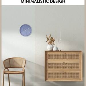 May include: A light blue wall clock with black hands and Roman numerals. The clock is hanging on a white wall. A wooden chair with a woven seat and back is in the foreground. A wooden dresser with three drawers and woven fronts is on the wall. A vase with dried wheat stalks is on top of the dresser.