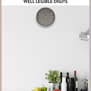 Op de afbeelding: Een grijze wandklok met zwarte cijfers en wijzers, hangend in een keuken met een wit aanrecht. Er staan groene kruiden in een metalen blik, rode en gele paprika's en flessen wijn en olie op het aanrecht.