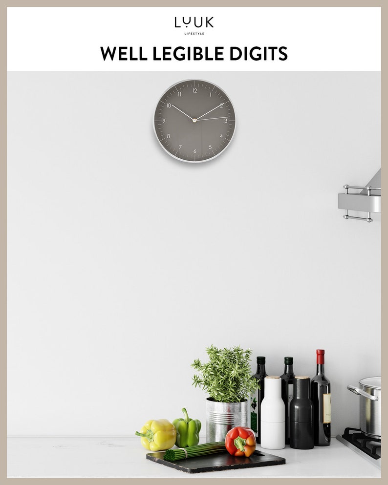 Op de afbeelding: Een grijze wandklok met zwarte cijfers en wijzers, hangend in een keuken met een wit aanrecht. Er staan groene kruiden in een metalen blik, rode en gele paprika's en flessen wijn en olie op het aanrecht.