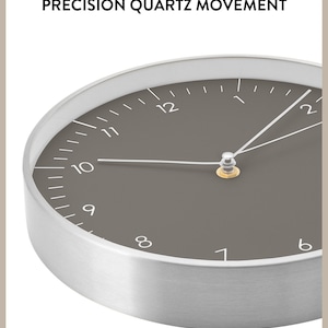 Op de afbeelding: Een close-up van een zilveren en grijze analoge klok met een donkergrijze wijzerplaat en witte markeringen. De wijzers van de klok zijn zilver en de wijzerplaat van de klok geeft "PRECISION QUARTZ MOVEMENT" aan.