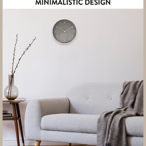 Op de afbeelding: Een grijze wandklok met witte wijzers en cijfers, hangend aan een witte muur boven een lichtgrijze bank met een bruin wollen deken. De bank staat in een woonkamer met een houten vloer en een klein bijzettafeltje met een vaas met gedroogde takken.