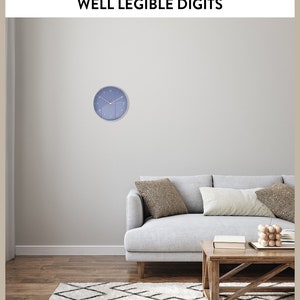 May include: A blue wall clock with white numbers and hands, hanging on a light gray wall above a gray sofa with two pillows. The sofa is in front of a wooden coffee table with a book and a wooden decorative object on top. The room has a white and black patterned rug on the floor.