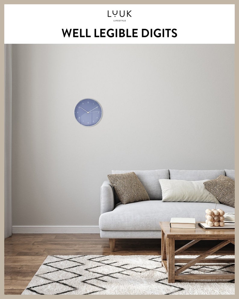 May include: A blue wall clock with white numbers and hands, hanging on a light gray wall above a gray sofa with two pillows. The sofa is in front of a wooden coffee table with a book and a wooden decorative object on top. The room has a white and black patterned rug on the floor.