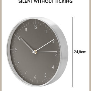 Op de afbeelding: Een ronde klok met een zilveren frame, een grijs gezicht en witte wijzers. De klok heeft een diameter van 24,8 cm en zegt "SILENT WITHOUT TICKING".