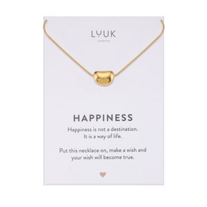 Könnte beinhalten: Goldkette mit einem kleinen, abgerundeten Anhänger, auf dem "Happiness" auf einer weißen Karte steht. Die Karte sagt auch "Happiness is not a destination. It is a way of life. Put this necklace on, make a wish and your wish will become true."