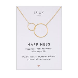 Könnte beinhalten: Goldkette mit zwei ineinander verschlungenen Kreisen, auf einer weißen Karte mit dem Text "LOUK LIFESTYLE" und "HAPPINESS" mit dem Zitat "Glück ist kein Ziel. Es ist eine Lebenseinstellung. Legen Sie diese Kette an, wünschen Sie sich etwas und Ihr Wunsch wird wahr."