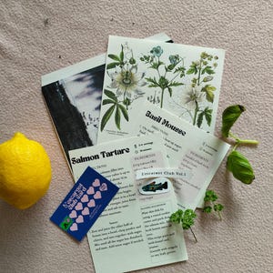 May include: A collection of recipe cards, a yellow lemon, and fresh herbs arranged on a pink surface. The cards detail recipes for Salmon Tartare and Basil Mousse. A small blue card with pink hearts and the text "L'escargot Club Card" is also present.