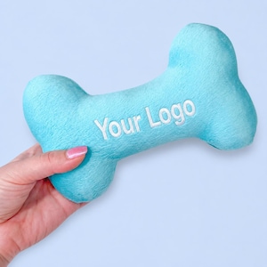 May include: A light blue plush dog toy shaped like a bone with the text Your Logo embroidered in white thread across the center. The fabric has a soft, fuzzy texture and the object features a classic bone silhouette with rounded ends.