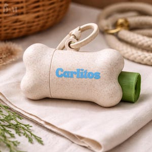 May include: Bone-shaped beige pet waste bag dispenser with the name "Carlitos" in blue. The dispenser has a silver clip and a roll of green bags is visible. The dispenser is on a beige cloth.