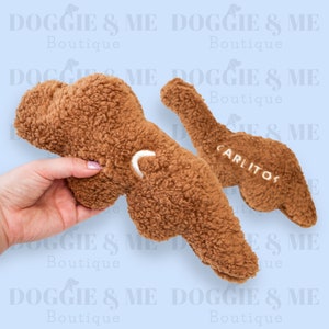 May include: Two brown plush dog toys, one held by a hand. The toys are shaped like a bone with a loop and the word "CARLITOS" embroidered on one. The background has the text "DOGGIE & ME Boutique".