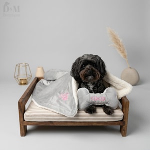 May include: A small black dog sits on a wooden pet bed with a striped cushion. The dog is wrapped in a gray fleece blanket with a white sherpa lining, featuring a pink paw print and a gray bone-shaped toy embroidered with the name COCO in pink.