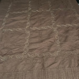 Puede incluir: Una colcha acolchada marr&oacute;n con un patr&oacute;n decorativo de remolinos y l&iacute;neas en un color marr&oacute;n m&aacute;s claro. La colcha tiene un patr&oacute;n de cuadr&iacute;cula de nueve cuadrados con un borde decorativo alrededor de cada cuadrado.