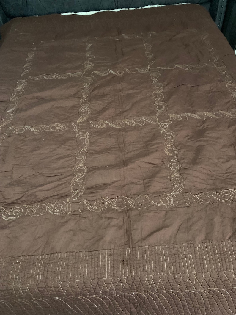 Puede incluir: Una colcha acolchada marr&oacute;n con un patr&oacute;n decorativo de remolinos y l&iacute;neas en un color marr&oacute;n m&aacute;s claro. La colcha tiene un patr&oacute;n de cuadr&iacute;cula de nueve cuadrados con un borde decorativo alrededor de cada cuadrado.