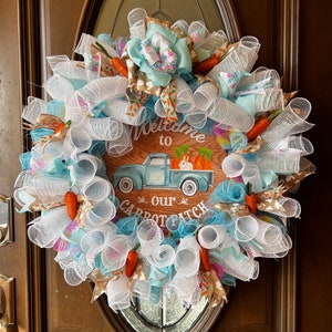 Puede incluir: Una corona decorativa de Pascua con un letrero de madera que dice "Welcome to our Carrot Patch". La corona presenta malla blanca, cintas azules y zanahorias artificiales. Un camión azul claro transporta zanahorias y un conejo.