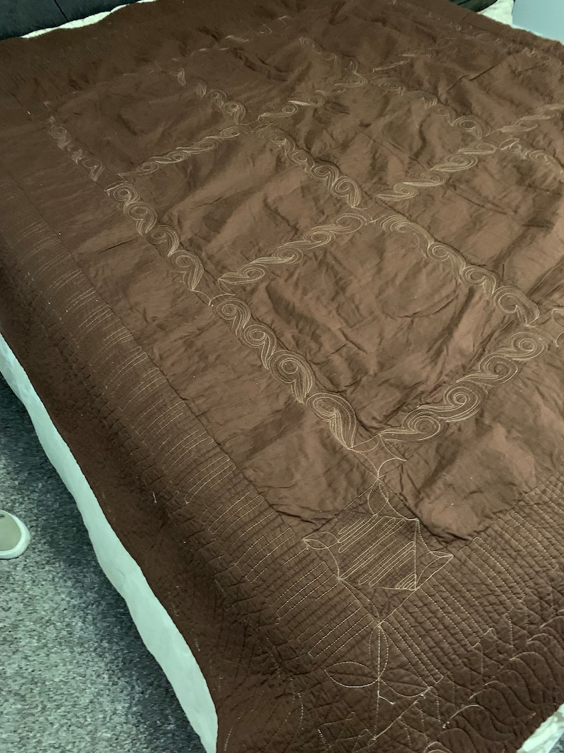 Puede incluir: Una manta de tiro acolchada marr&oacute;n con un patr&oacute;n de puntadas decorativas. La manta tiene un brillo sutil y una textura suave.