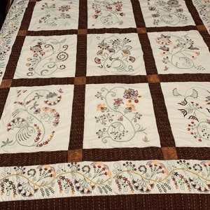 Puede incluir: Una colcha blanca con un borde marr&oacute;n y un patr&oacute;n repetitivo de dise&ntilde;os florales bordados en tonos marrones, verdes y blancos.