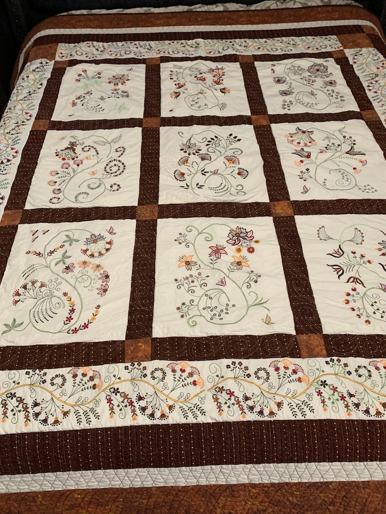 Puede incluir: Una colcha blanca con un borde marr&oacute;n y un patr&oacute;n repetitivo de dise&ntilde;os florales bordados en tonos marrones, verdes y blancos.