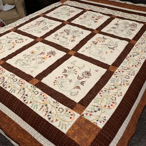 Puede incluir: Una colcha blanca con un borde marr&oacute;n y bordados florales intrincados en tonos de marr&oacute;n, verde y naranja. La colcha est&aacute; compuesta por nueve cuadrados, cada uno con un dise&ntilde;o floral diferente.