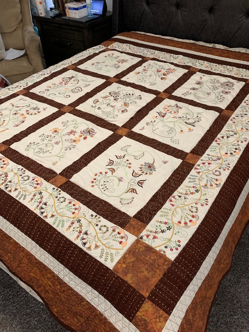 Puede incluir: Una colcha blanca con un borde marr&oacute;n y bordados florales intrincados en tonos de marr&oacute;n, verde y naranja. La colcha est&aacute; compuesta por nueve cuadrados, cada uno con un dise&ntilde;o floral diferente.