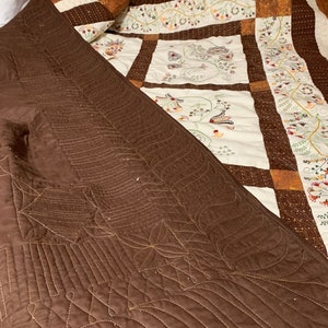 Puede incluir: Una manta acolchada marr&oacute;n con un patr&oacute;n decorativo de costuras. La manta est&aacute; doblada sobre una colcha blanca y marr&oacute;n con dise&ntilde;os florales bordados.
